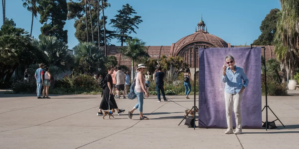 Alice raises a hand to her brow in a breezy salute, dressed in a light blue shirt and cream pants, at a purple backdrop on the lively Balboa Park promenade.