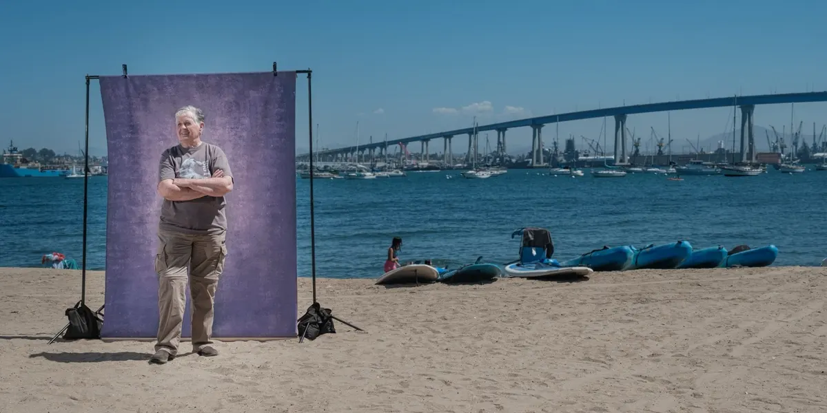 Arlene stands with arms crossed and gaze lifted toward the water, in a gray tee and khaki cargo pants, at a purple backdrop on the beach with the Coronado Bridge in the distance.