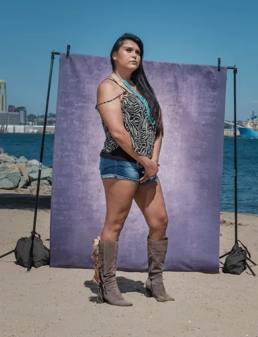 Camie gazes into the distance in an animal-print top and tall suede boots, standing at a purple backdrop on the sand.