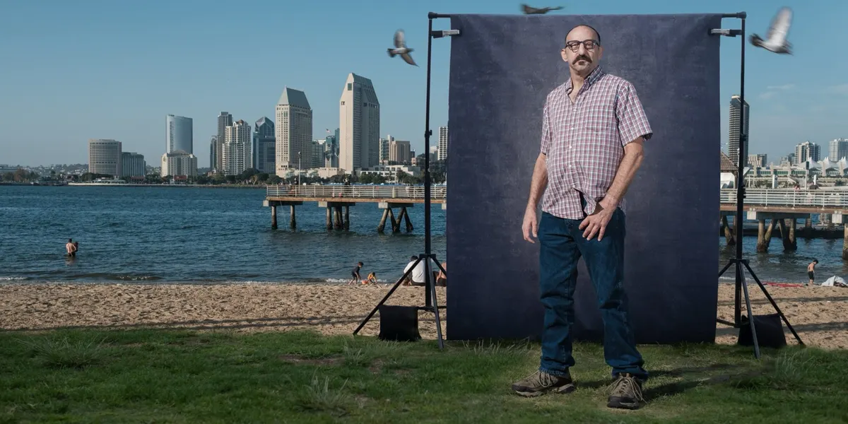 Jeffrey stands steady in a plaid shirt and jeans as pigeons take flight around him, posed at a navy backdrop on the Coronado shoreline with the San Diego skyline behind him.