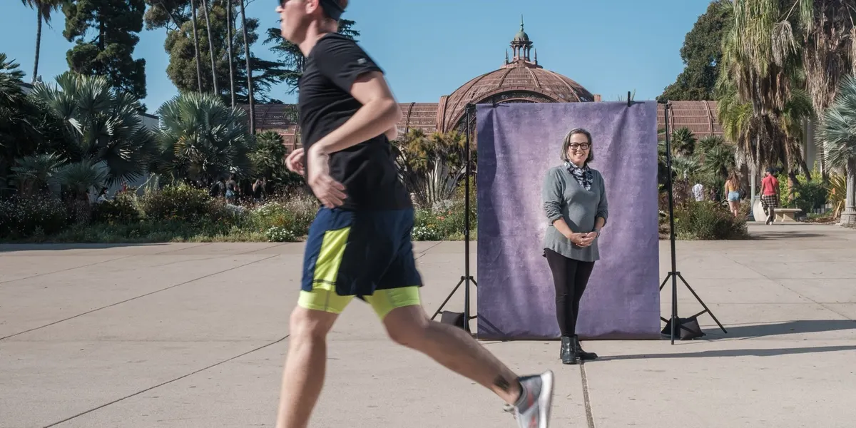 Linda smiles softly in a cozy gray sweater and scarf at a purple backdrop on the Balboa Park promenade, while a jogger blurs past in the foreground.