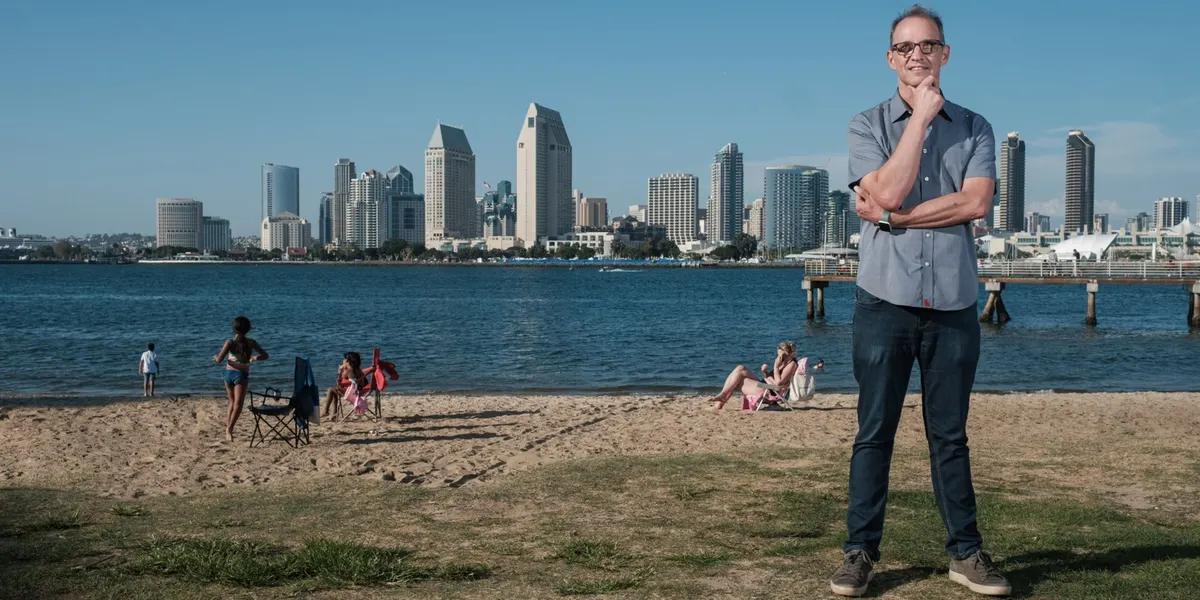 Michael rests his chin on his hand with a warm smile, wearing a gray button-up, standing on a grassy bank with the beach and San Diego skyline glowing behind him.