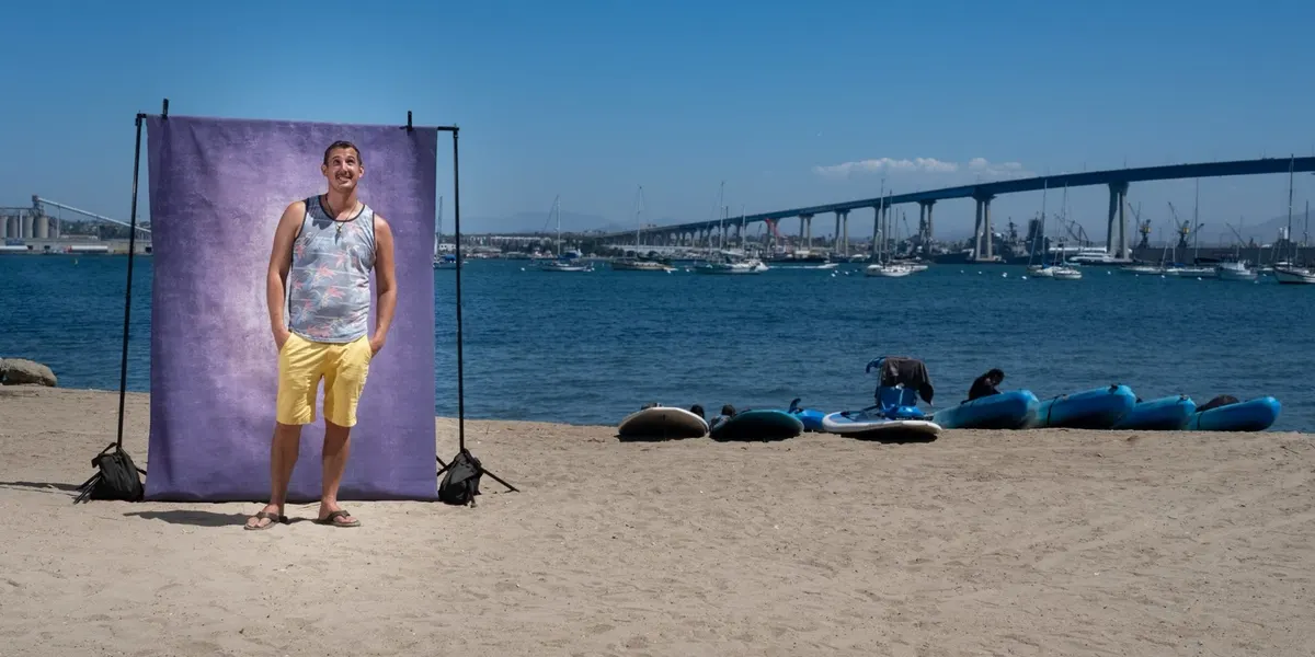 Michael looks up with a bright smile in a floral tank top and yellow shorts, standing at a purple backdrop on the beach with the Coronado Bridge behind him.