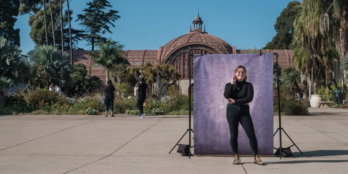 Miki smiles warmly in a black turtleneck, one hand raised to their cheek, standing at a purple backdrop on the sunlit Balboa Park plaza.