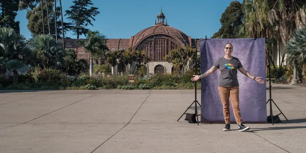 Mollie throws her arms open wide in a gray tee with a rainbow graphic, grinning in front of a purple backdrop with the Botanical Building and gardens behind her.