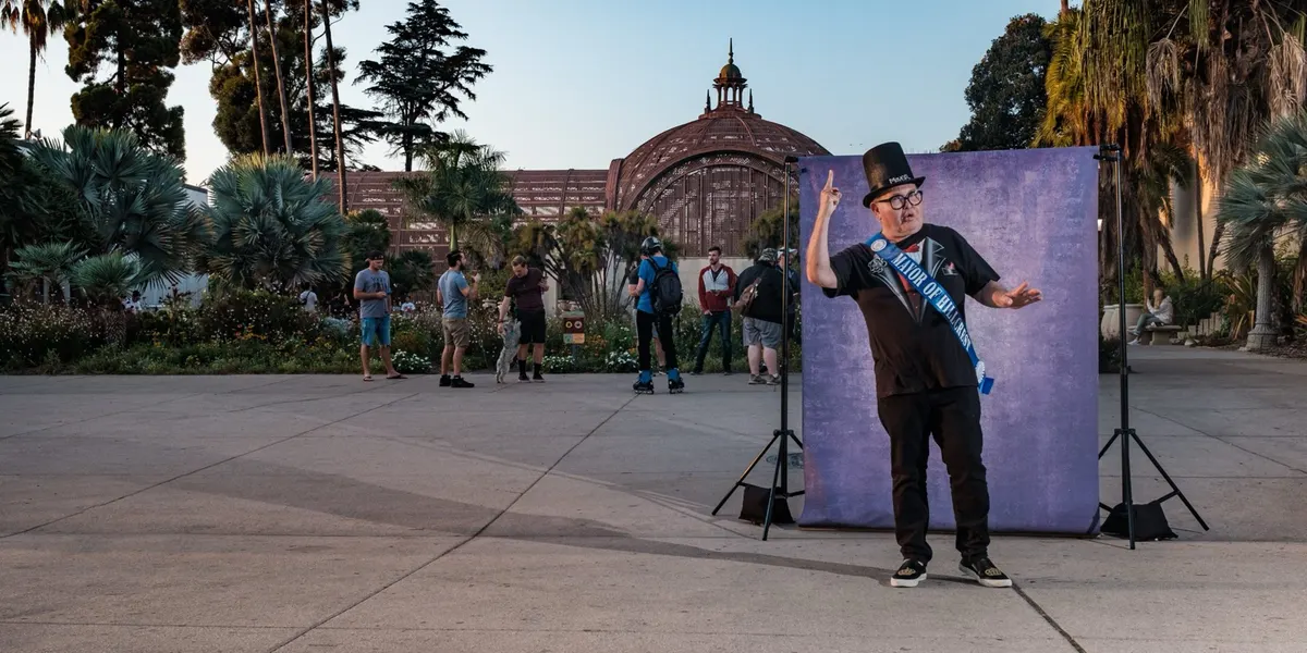 Nicole points skyward in a top hat and a celebratory sash over a black outfit, mid-performance in front of a purple backdrop on the Balboa Park promenade at dusk.