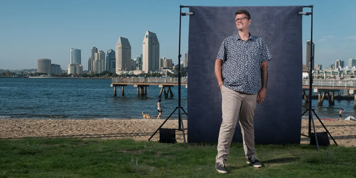 Tony smiles off to the side in a blue patterned shirt, posed before a navy backdrop on the Coronado shoreline with the San Diego skyline behind him.
