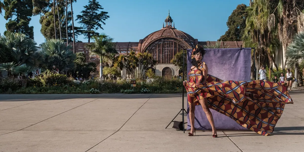 Tootie poses in a sculpted headpiece and heels as a sweeping fan-print gown billows out beside her, in front of a purple backdrop near the Balboa Park Botanical Building.