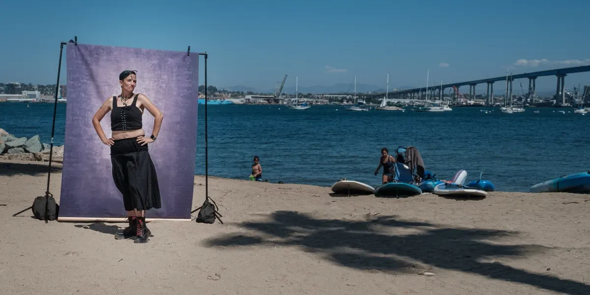 A community member stands hands-on-hips in a black corset top and lace-up boots, posed at a purple backdrop on the sand with the Coronado Bridge in the distance.